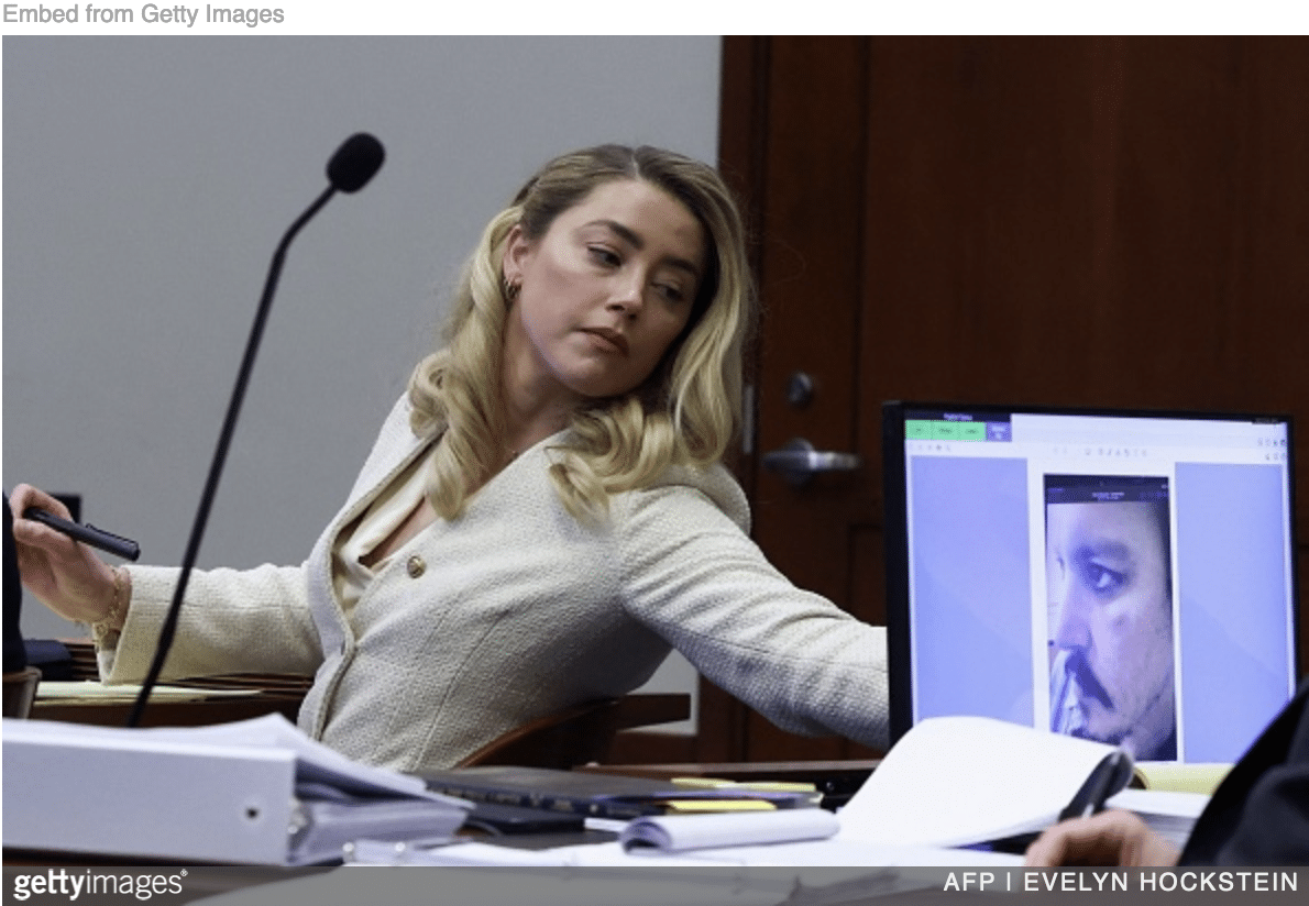 Amber Heard testifying at defamation trial with image of Depp on small monitor beside her.